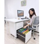 A professional lady seated in an office chair reaching into the bottom filing drawer of a white iSimply steel mobile pedestal to retrieve document folders.