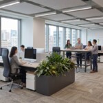 A wide-angle photo of multiple black Vero PU leather high-back chairs used across dynamic workstations in a bustling, collaborative open-plan co-working office, with teams visible in the background and diverse city views.