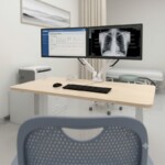 A white FlexiSpot dual monitor arm securely clamped to a desk in a bright doctor's consultation room, holding two screens for efficient medical record management.